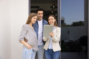 Positive professional realtor woman meeting with young couple of renters at apartment, using tablet, showing screen to customers, standing on rental doorstep, talking , laughing