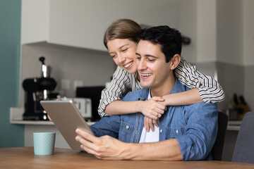 Cheerful attractive Hispanic couple using tablet computer, watching online content, activating kitchen appliance with smart home virtual assistant, hugging, smiling, laughing