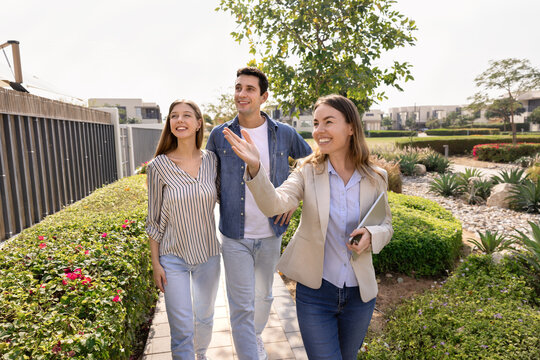 Happy professional realtor woman giving tour around well suburb neighborhood for house buyers, showing apartment exterior, location to young couple of customers, walking outside, pointing hand away