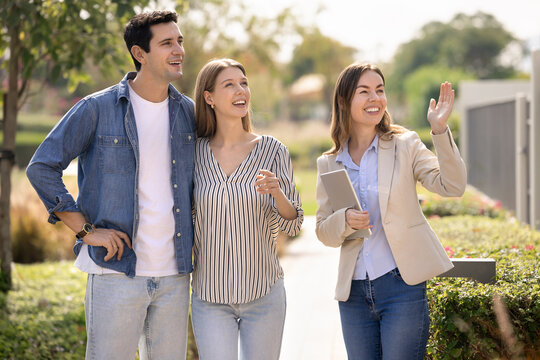 Happy young Hispanic couple and real estate agent viewing good area for living, looking away at new house, discussing rent, property buying, mortgage. Realtor showing neighborhood to customers