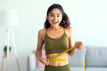 Portrait of young Indian woman measuring her waist with tape indoors, smiling at camera. Beautiful...