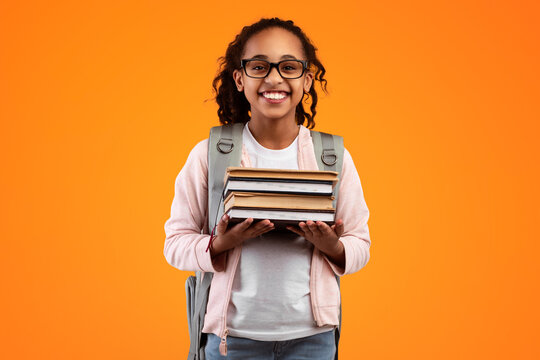 Education Concept. Portrait of smiling African American student girl holding stack of academic books and looking at camera, wearing eyeglasses and backpack, yellow color studio background