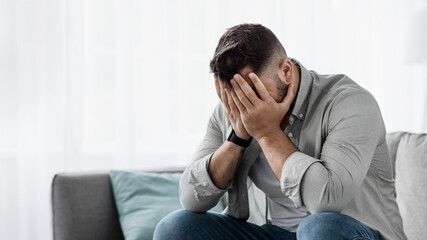 Shame despair, portrait of sad man suffers from headache, stress, depression. Failure unhappy millennial european handsome guy crying, covering face with hands, sit on sofa in living room interior