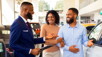 Happy Black Couple Talking To Salesman While Choosing New Car In Dealership Center, Smiling African American Spouses Purchasing Family Vehicle In Modern Automobile Showroom, Closeup Shot
