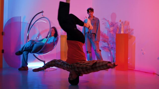 Happy hipster doing head spin while friend looking at performance with neon light dancing studio. Professional caucasian man moving to music while smart people sitting and pose at camera. Regalement.