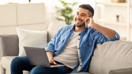 Portrait of smiling young jew man in yarmulke talking on mobile phone, working remotely sitting on comfortable couch with laptop at home office. Positive guy making business call, blurred background