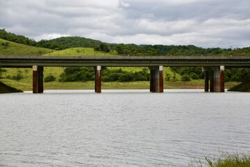 Ponte de concreto em cima de uma represa com movimento de carros sobre ela e fundo vibrante da natureza.