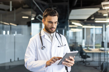 Serious thinking male doctor working inside clinic office with tablet computer. Doctor using app...
