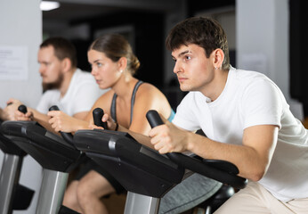 Obraz premium In gym, guy does exercises using stationary bicycle. Young group of athletes trains in gym. Physical education and sports, active healthy lifestyle.