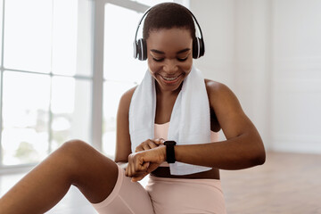 Smiling young African American woman looking at activity fitness tracker during workout break, checking heart rate and burned calories. Happy lady wearing wireless headphones, sports bra and towel