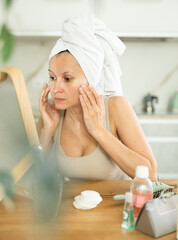 Adult woman with towel wrapped around head meticulously checking face in mirror during skincare...
