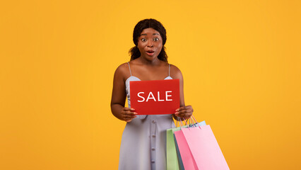 Surprised millennial black woman holding shopping bags and SALE sign over orange studio background....