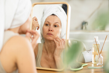 Adult woman with towel wrapped around head meticulously checking face in mirror during skincare...