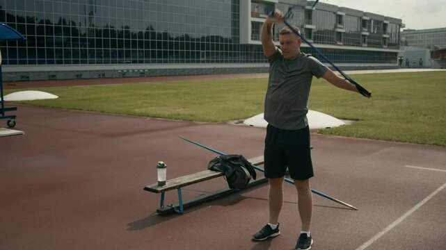 Full shot of young Caucasian male javelin thrower with missing hand performing warmup routine at outdoor stadium &ndash; standing on grass, repeating shoulder joint mobility exercises with elastic band