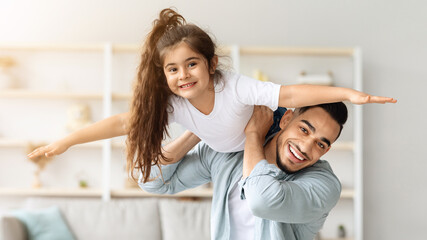 Fototapeta premium Happy arab father and daughter having fun together at home, middle-eastern young man dad playing airplane with his cute daughter in living room, lifting kid up, holding on shoulder, copy space