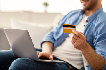 Closeup Cropped View Of Unrecognizable Guy Using Laptop And Credit Card At Home For Online Payments, Sitting On Couch, Transferring Money, Shopping In Internet Or Booking Vacation, Selective Focus