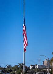 A large United States flag at half mast against a blue sky. There is very little wind so the flag...