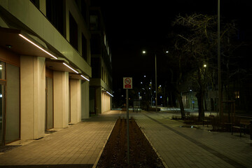 spacious pedestrian space at night with a flowerbed in the middle and a technological sign, illuminated by extended lighting on the building