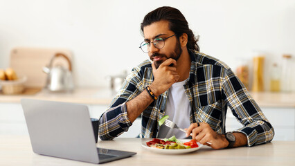 Pensive indian man freelancer having snack while working from home during coronavirus pandemic,...