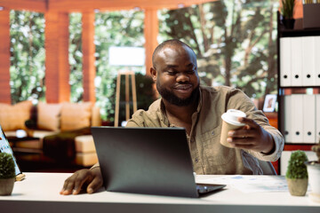 Business owner working remotely on a laptop and drinking coffee, enjoying his home based setup for enterprise activity. Handling operations online, showcasing successful telecommuting.