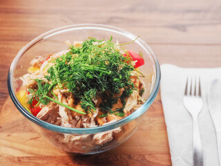 Fresh salad with tuna, tomato and dill in a glass round bowl standing on a wooden board and table. Easy healthy portion of food.