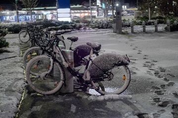 A bike is parked next to a bike hire rack. The bike is covered in snow and the snow is piled up on the ground. The scene is quiet and peaceful, with the snow covering the ground and the bike