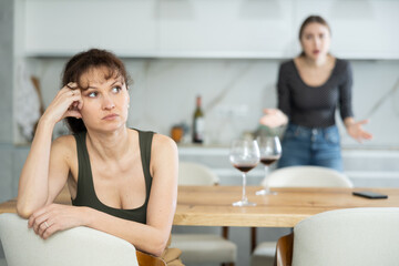Sad middle-aged woman sitting at table and other woman brawling to her behind in the kitchen