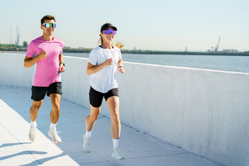 Couple jogging along a waterfront path on a sunny day wearing colorful sunglasses and sport attire