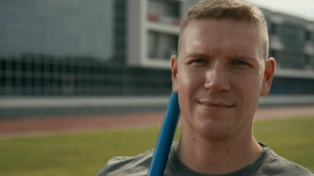 Closeup portrait of face of young professional Caucasian male javelin thrower standing at outdoor stadium during workout, looking at camera and smiling confidently, while preparing for championship
