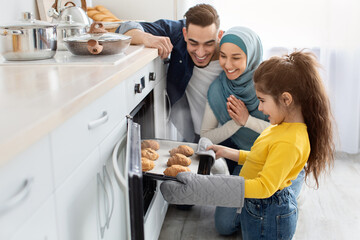 Happy muslim family of three baking together in kitchen, cute little daughter taking tray with fresh baked croissants out of oven, cheerful middle eastern parents and child enjoying homemade pastry