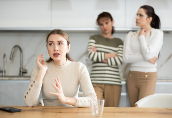 Offended middle-aged woman sitting in the kitchen while two women talking angrily to her