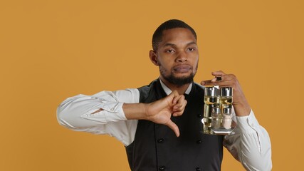 Restaurant staff giving a thumbs down sign against yellow background, carrying a cruet set with essential condiments at tables. Waiter in customer service industry, dislike symbol. Camera A.