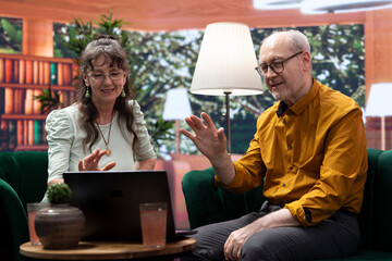 Elderly man and woman waving at grandchildren on online videocall, maintaining the long distance...