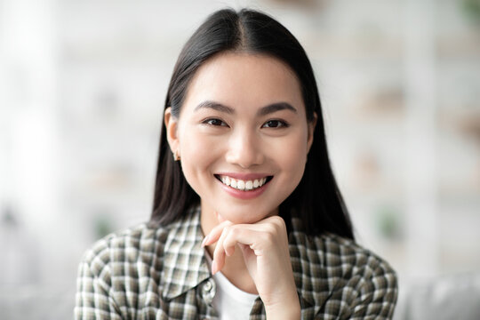 Closeup potrait of attractive long-haired asian woman in cozy outfit chilling at home, smiling at camera, copy space. Home interior, millennials lifestyle, relaxation and leisure concept