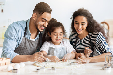 Positive arab father, mother and daughter making biscuits together, cutting pastry dough, middle-eastern mom and dad showing their little kid how to make cookies, wearing aprons, kitchen interior