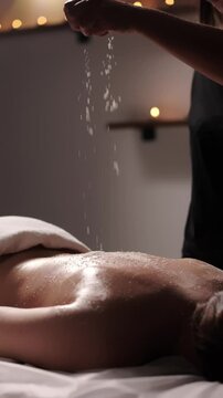Hand of masseuse gently sprinkles coconut scrub onto a woman back before massage in spa salon in backlight. Perfect concept for promoting body care, relaxation, wellness. Vertical shots, advertising
