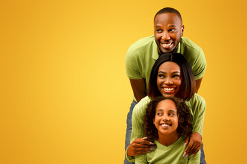 Fototapeta premium Promo concept. Happy black family of three looking aside at free copy space on yellow studio background, standing behind each other. Empty space for dvertisement