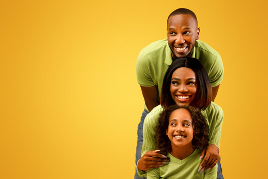 Promo concept. Happy black family of three looking aside at free copy space on yellow studio background, standing behind each other. Empty space for dvertisement