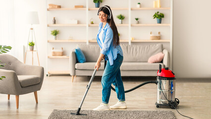 Cleaning With Pleasure. Happy young woman vacuuming rug floor carpet using hoover, listening to...