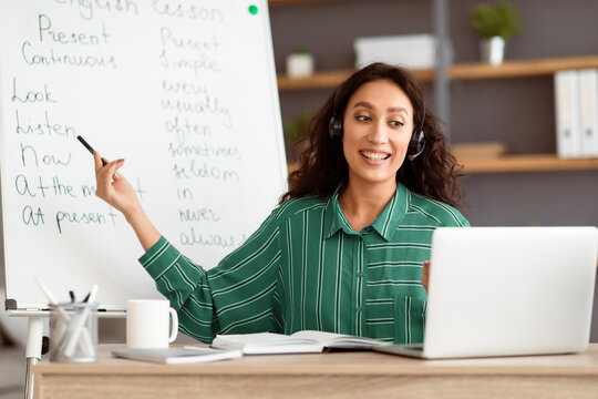 Elearning Concept. Portrait of smiling lady teacher having video conference call using laptop. Lady in wireless headphones talking to webcam, recording tutorial on pc for students pointing at board