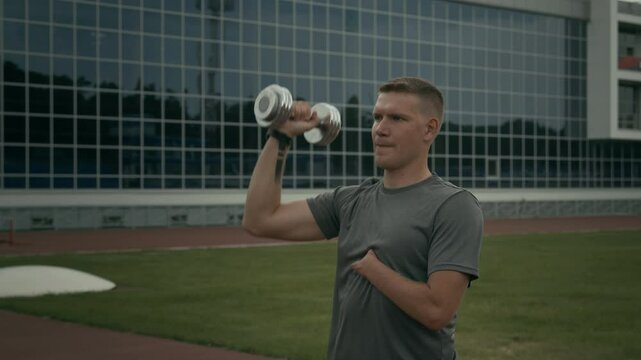 Medium shot of Caucasian male para athlete with limb difference disability, missing hand, performing shoulder press exercise with dumbbell at outdoor stadium in summer, looking ahead
