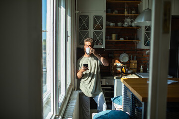 Man drinking coffee and using smartphone in sunny kitchen