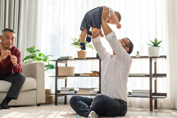 Joyful moment of multigenerational family at home, father holding up his baby smiling and playing...