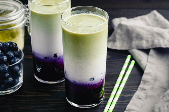 Iced Blueberry Matcha Lattes in Tall Glasses: Cold lattes made of layered green tea, milk, and blueberries with paper straws and ingredients nearby