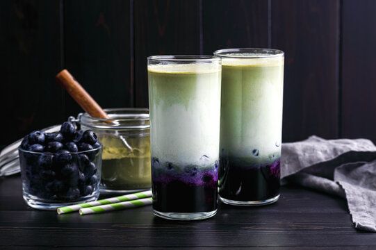 Iced Blueberry Matcha Lattes in Tall Glasses: Cold lattes made of layered green tea, milk, and blueberries with paper straws and ingredients nearby