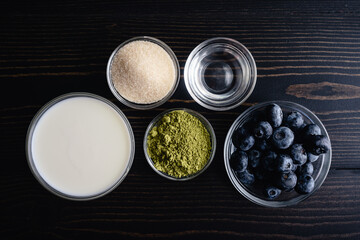 Blueberry Matcha Latte Ingredients on a Dark Wooden Table: Green tea powder, fresh blueberries, organic sugar, milk, and water in glass prep bowls