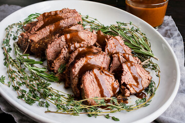 Sliced Tri-Tip Roast Beef with Red Wine Glaze: Slices of roast beef with sauce surrounded with sprigs of thyme and rosemary on a ceramic plate