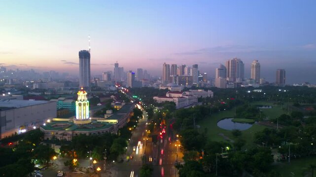 Aerial: An Illuminated City Skyline Glows At Dusk With Prominent Buildings, A Lighted Tower, And Busy Streets Under A Pastel Sky - Manila, Philippines