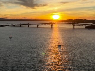 Obraz premium Golden sunset over the bridge, reflecting on the water. A sailboat and other small boats are visible on the water. Auckland Harbour Bridge, Auckland, New Zealand