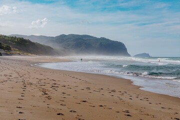 Tranquil beach scene with gentle waves, footprints in the sand, and a surfer walking along the shore. Coastal serenity. Opoutere, Whangamata, Coromandel Peninsula, New Zealand